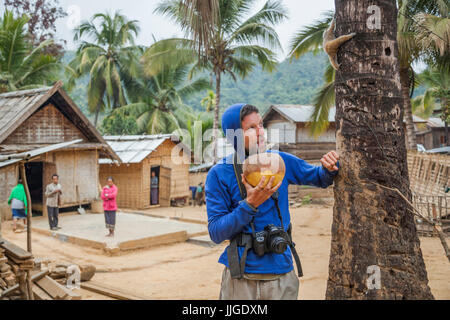 Robert Hahn gorgées sur une noix de coco alors qu'un slow loris Nycticebus (sp.), capturés dans la forêt environnante et gardé comme animal de compagnie, monte dans un cocotier en interdiction a Dan, Laos. Banque D'Images