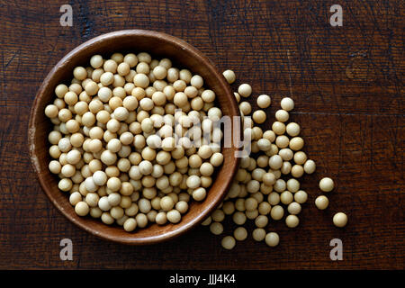 Sécher les graines de soja dans un bol en bois isolé sur bois marron foncé à partir de ci-dessus. Soja déversé. Banque D'Images