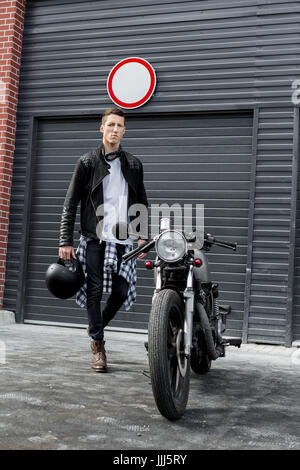 Beau jeune mec en noir veste biker bottes et aller à son style classique cafe racer motorcycle portes industrielles comme arrière-plan. Ma coutume de vélo Banque D'Images