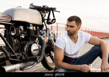 Beau happy rider guy avec barbe et moustache s'asseoir près de son style classique biker moto cafe racer et regarder vers le côté. Fait sur commande de vélo en vint Banque D'Images