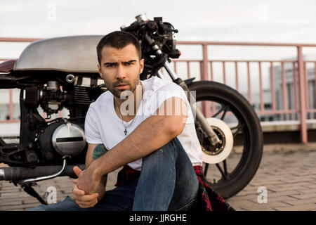 Beau happy rider guy avec barbe et moustache s'asseoir près de son style classique biker moto cafe racer et regarder vers l'appareil photo. Fait sur commande de vélo de vi Banque D'Images