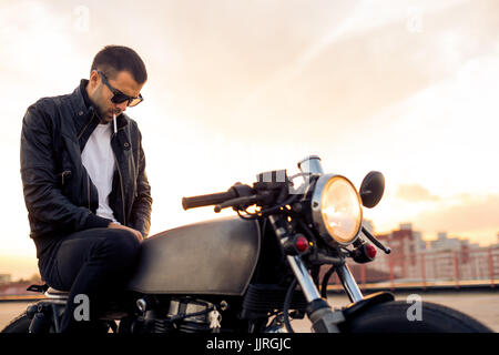 Handsome Guy Rider avec barbe et moustache en noir Veste motard et lunettes de mode 1900 fumée et s'asseoir sur le style classique du cafe racer sur moto Banque D'Images