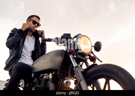 Handsome Guy Rider avec barbe et moustache en noir lunettes fashion 1900 fumeurs et corriger biker jacket s'asseoir sur un style classique du cafe racer motorb Banque D'Images