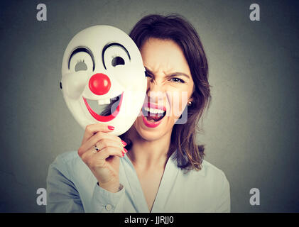 Hurlant de colère Portrait woman taking off masque clown heureux isolé sur fond de mur gris. Les émotions humaines sentiments négatifs Banque D'Images