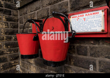 Voir de 2 vieux seaux d'incendie rouge & noir accroché sur la paroi de la plate-forme à la gare historique, pittoresque Oakworth - Keighley & Worth Valley Railway, England, UK. Banque D'Images