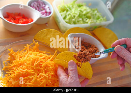 Un homme fait des tacos à la maison avec des coquilles à taco, boeuf haché, laitue, oignons, tomates et fromage râpé Banque D'Images