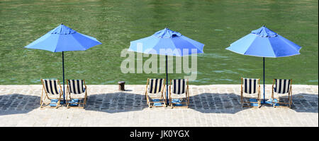 Trois parasols bleu rayé bleu et blanc avec des chaises longues au soleil sur le quai de la Seine avec l'eau verte. Banque D'Images