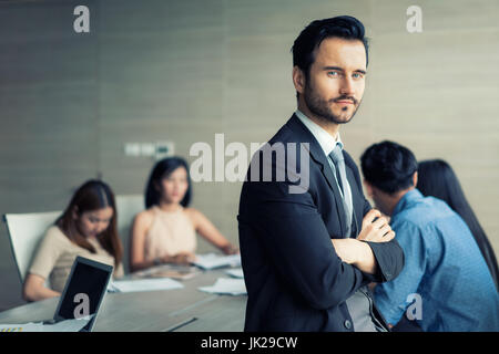 Happy business man standing with arms crossed au bureau pendant que ses collègues discutant en arrière-plan. Banque D'Images