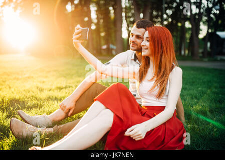 Love couple fait sur téléphone appareil selfies en parc d'été sur le coucher du soleil. Date romantique de jolie femme et jeune homme Banque D'Images