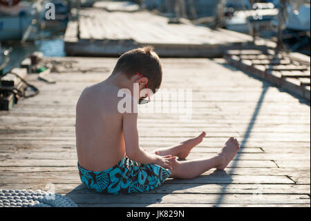 Petit enfant portant des shorts et lunettes de soleil assis sur la jetée en bois en journée ensoleillée Banque D'Images