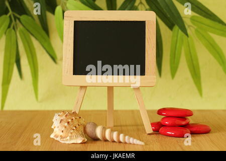 Vide petit tableau noir à écrire un message posé sur un chevalet sur un sol en bois avec plage de galets et de rouge sur un fond de feuillage vert Banque D'Images