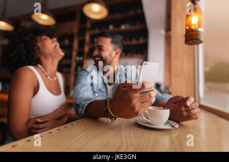 Young couple at coffee shop. Man holding mobile phone avec laughing woman sitting by. Banque D'Images