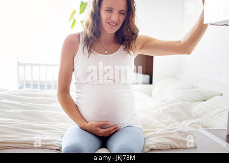 Belle Jeune femme enceinte, dans la douleur avec la contraction du travail, assis sur le lit dans la chambre, prepraring pour donner une naissance Banque D'Images