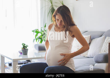 Jeune femme enceinte, avoir contraction douloureuse, en commençant le travail, assis sur le canapé Banque D'Images