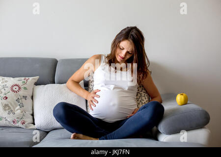 Jeune femme enceinte, avoir contraction douloureuse, en commençant le travail, assis sur le canapé Banque D'Images