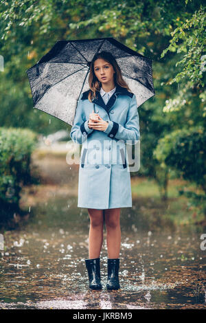 Portrait of teenage girl holding umbrella en se tenant sur le chemin au milieu des arbres dans la pluie Banque D'Images