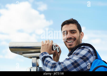 Portrait of young handsome man smiling at camera while heureusement à l'aide de jumelles à monnayeur sur plate-forme d'observation sur le toit bleu clair contre Banque D'Images