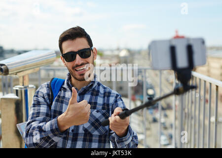 Portrait de beau jeune homme prenant photo selfies en se tenant sur le toit contre une vue panoramique sur la ville et les jumelles dans l'arrière-plan Banque D'Images