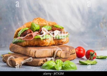 Croissant sandwich avec de la viande fumée jambon de Parme, tomates séchées, épinards frais et basilic sur fond gris texturé en pierre Banque D'Images