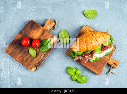 Croissant sandwich avec de la viande fumée jambon de Parme, tomates séchées, épinards frais et le basilic sur les petites planches de bois rustique en pierre sur fond gris texturé Banque D'Images