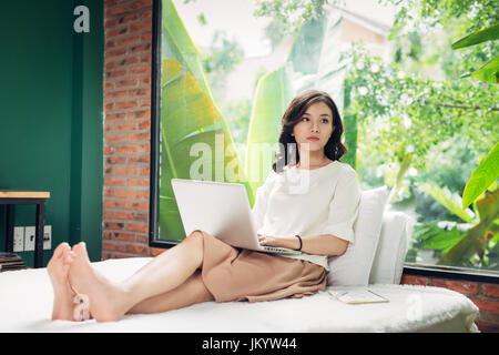 Beau young smiling woman working on laptop while sitting on bed at home près de grande fenêtre. Banque D'Images