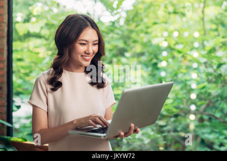 Beau young smiling woman working on laptop while sitting on bed at home près de grande fenêtre. Banque D'Images