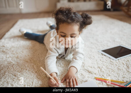 Adorable petit african american girl dessin avec crayons et couché sur un tapis à la maison Banque D'Images