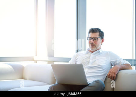 Beau et mature portant des lunettes concepteur, assis confortablement sur un canapé tout en travaillant sur son ordinateur portable Banque D'Images