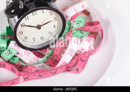 Couleur emmêlés les rubans à mesurer et classic noir réveil sur la plaque sur fond blanc, symbolisant le temps de manger ou de remise en forme régime perdre du poids Banque D'Images