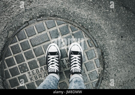 Saint-pétersbourg, Russie - 30 mai 2017 : les pieds dans une paire de toile noir Chuck Taylor All-Stars chaussures occasionnels sont sur un couvercle de trou d'urbain Banque D'Images