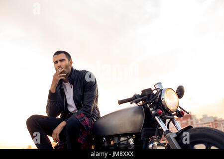 Handsome Guy Rider avec barbe et moustache de biker jacket fumeurs, de penser et de s'asseoir sur un style classique du cafe racer moto au coucher du soleil. Fait sur commande de vélo Banque D'Images