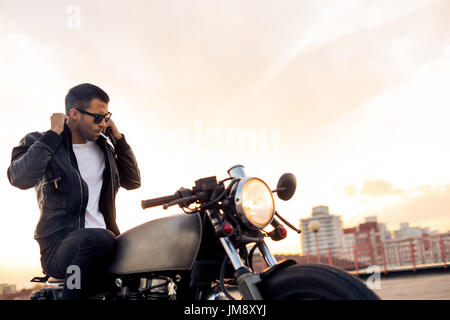 Handsome Guy Rider avec barbe et moustache 1900 fumeurs et corriger biker jacket s'asseoir sur un style classique du cafe racer moto au coucher du soleil. La coutume de vélo m Banque D'Images