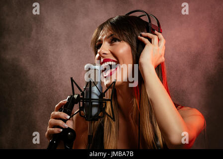 Portrait of a young woman singer avec des écouteurs à l'avant du microphone. Chanter avec la bouche grande ouverte et avec une expression de bonheur sur son visage Banque D'Images