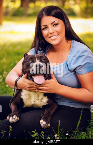 Belle jeune femme et son joli stafford terrier assis sur l'herbe dans le parc et d'apprécier. Regardant cemera. Banque D'Images