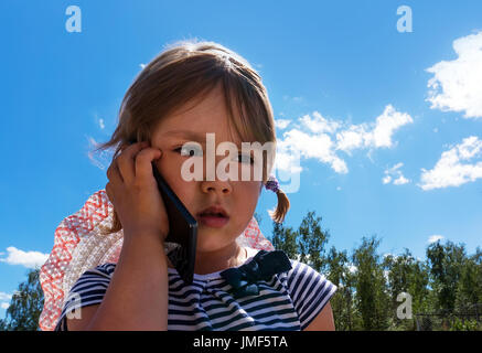 Petite fille avec un regard triste et malheureux yeux parler au téléphone avec ma mère sur le fond de ciel bleu à l'extérieur dans le parc. Banque D'Images