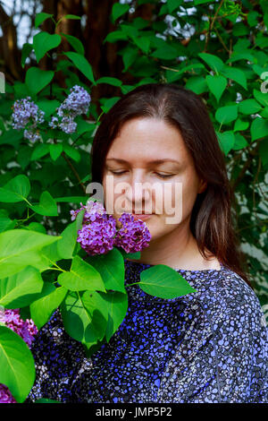 Girl offre une odeur de lilas. Lumière bouclés ci-dessous ses épaules, longue robe blanche d'été, chapeau avec panier de fleurs dans les mains est debout près de b Banque D'Images