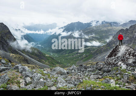 Jeune homme dans l'arrière de casquettes et rouge veste de pluie se trouve en face d'une immense vallée glaciaire avec des nuages de bas c'est les bords et les plantes vertes de remplir le Banque D'Images