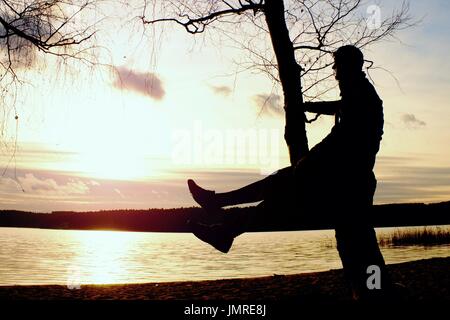 Sur l'homme arbre. Silhouette d'homme seul s'asseoir sur une branche du bouleau, en face de la coucher du soleil à rives. Banque D'Images