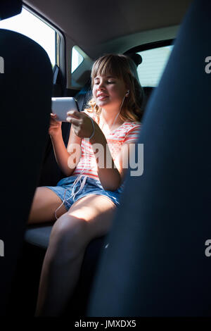 Teenage girl in headphones using mobile phone dans le siège arrière de voiture Banque D'Images