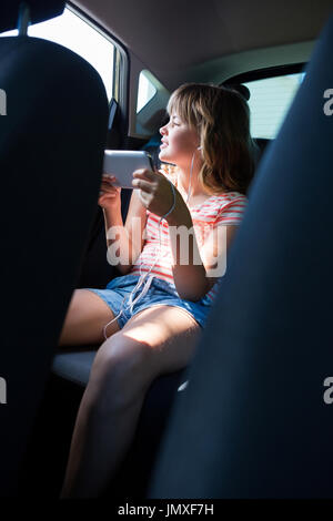 Teenage girl in headphones using mobile phone dans le siège arrière de voiture Banque D'Images