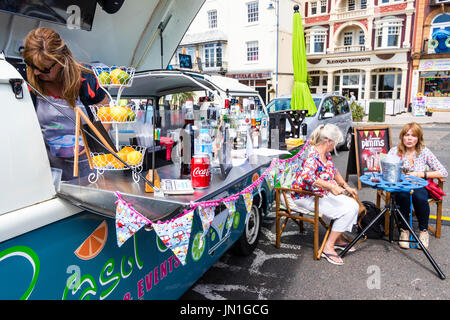 Le camping-car Volkswagen bleue modifiée en un bar mobile, mis en place avec des boissons et des bouteilles sur le comptoir sur le côté du van à l'extérieur lors de cas de Ramsgate en plein soleil. Deux femmes assises à table d'alcool à côté de van. Banque D'Images