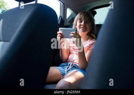 Teenage girl in headphones using mobile phone dans le siège arrière de voiture Banque D'Images