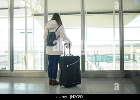 Fille avec une valise dans l'aéroport salle d'attente. Jeune passager regarde à travers une grande fenêtre sur la piste. Banque D'Images