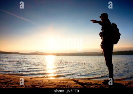 Homme heureux avec la main dans l'air. Grand randonneur dans les vêtements de sport sac à dos sombre avec des stands sur la plage admirer le coucher du soleil à l'horizon avec orange ciel rose avec c Banque D'Images