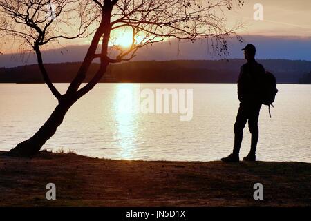 Homme heureux avec la main dans l'air. Grand randonneur dans les vêtements de sport sac à dos sombre avec des stands sur la plage admirer le coucher du soleil à l'horizon avec orange ciel rose avec c Banque D'Images