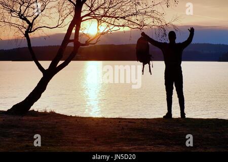 Homme heureux avec la main dans l'air. Grand randonneur dans les vêtements de sport sac à dos sombre avec des stands sur la plage admirer le coucher du soleil à l'horizon avec orange ciel rose avec c Banque D'Images