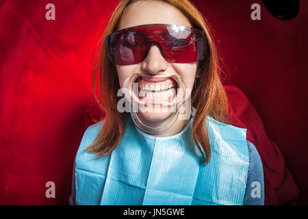 Jeune femme rousse à lunettes rouge avec porte-dents blanchissant des dents de dentiste, jusqu'à obtention d'une chaise design blanc sur rouge Banque D'Images