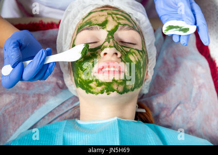 Un gros plan de l'esthéticienne en bleu à l'aide de gants spéciaux un pinceau de maquillage fait un masque de spa thérapeutiques d'algues bleues contre le vieillissement d'un jeune h-rouge Banque D'Images