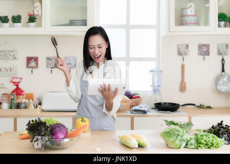 Jeune femme asiatique gaie est la cuisson dans la cuisine avec joie. Elle est debout et holding digital tablet de recette. Femme asiatique est de toucher un bois sp Banque D'Images