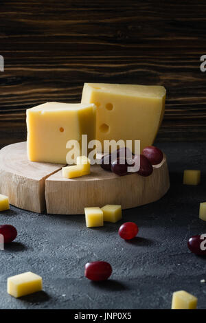 Disque suédois fromage jaune avec des trous coupé avec raisins rouges sur les tranches en bois sombre sur fond rustique Banque D'Images